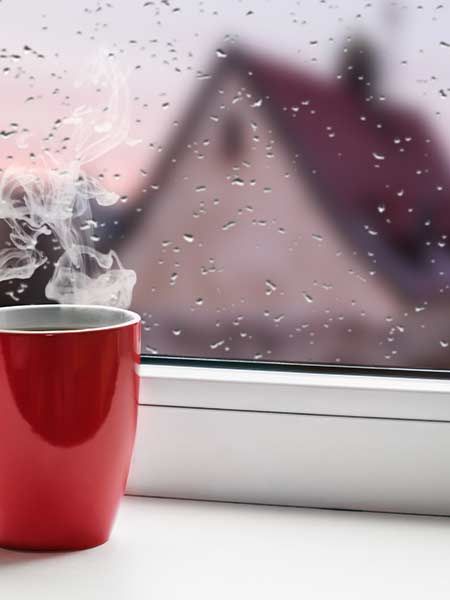 Steaming red mug on a windowsill with raindrops, blurred house in background.
