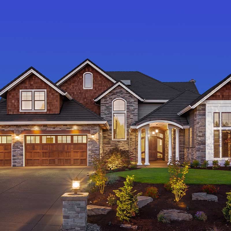 Two-story house with stone and wood exterior, brown garage doors, and landscaped yard lit at dusk.