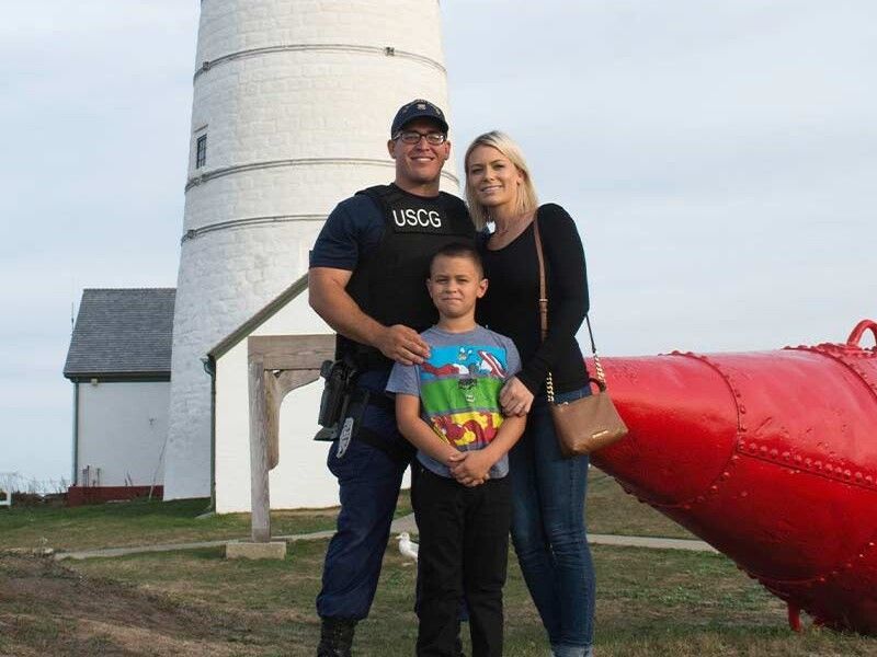 Family posing in front of a white lighthouse and red buoy. The person on the left is wearing a Coast Guard uniform.