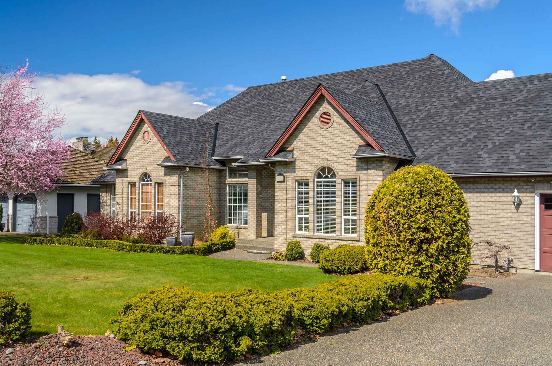 Beige brick house with gray roof, green lawn, and flowering tree under a blue sky.