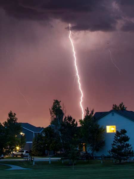 Lightning strike illuminates a suburban neighborhood at dusk. Dark clouds, purple sky, and a house with lit window.