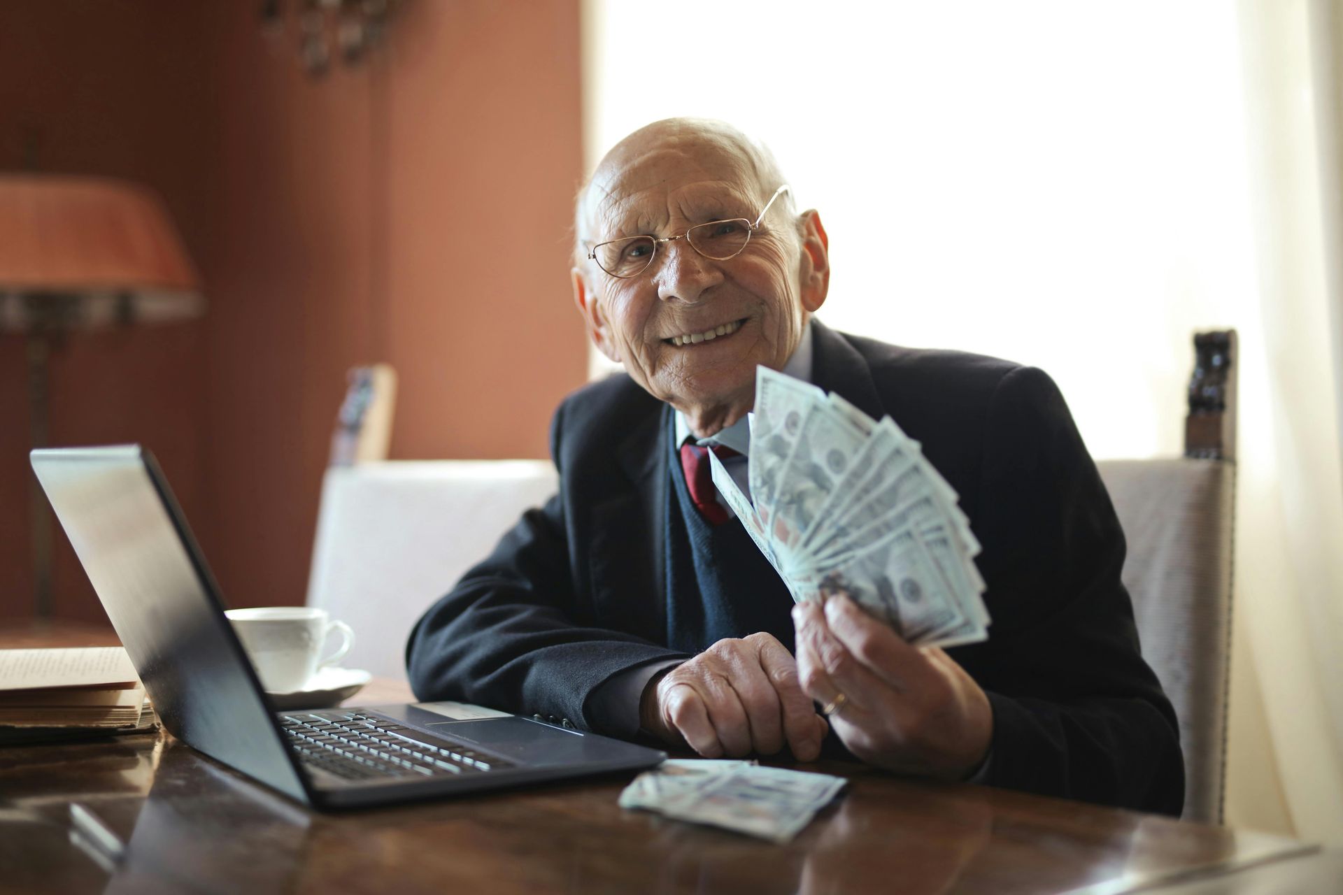 An elderly man is sitting at a table holding a fan of money.