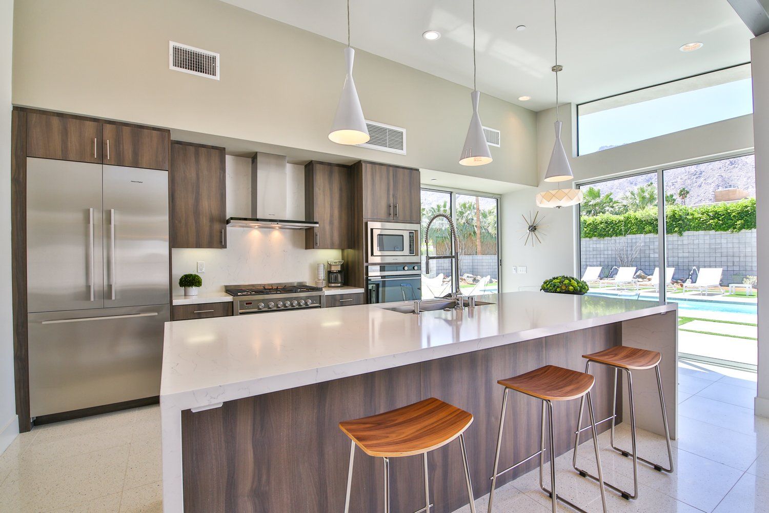 A kitchen with a large island and stools and a swimming pool in the background.