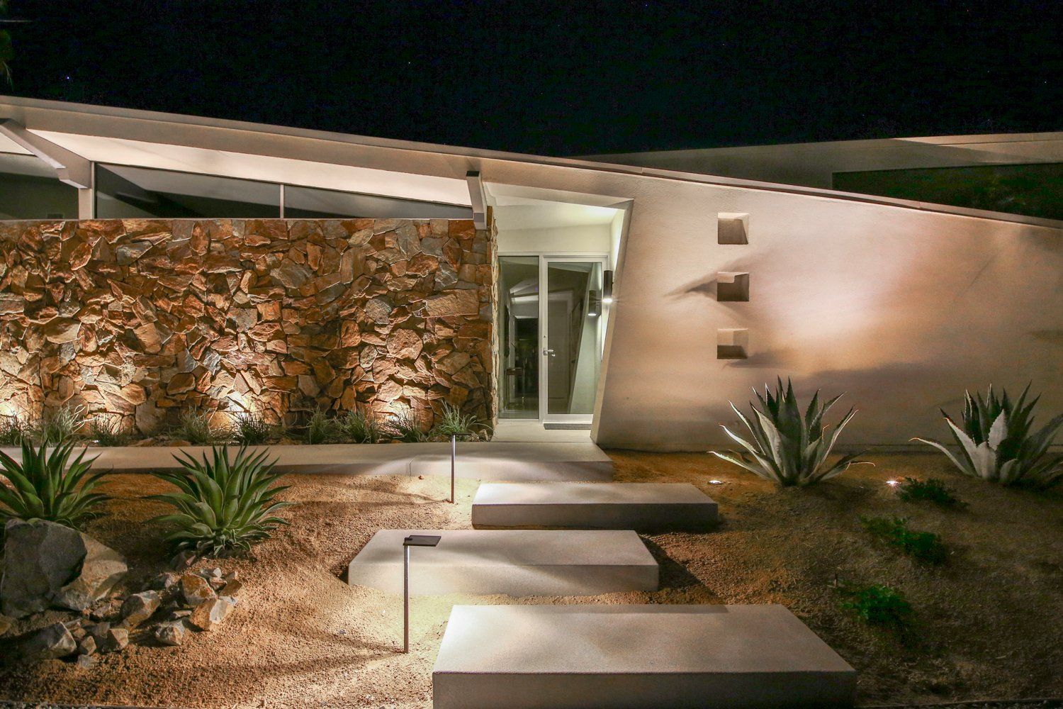 A modern house with a stone wall and steps leading to it at night.