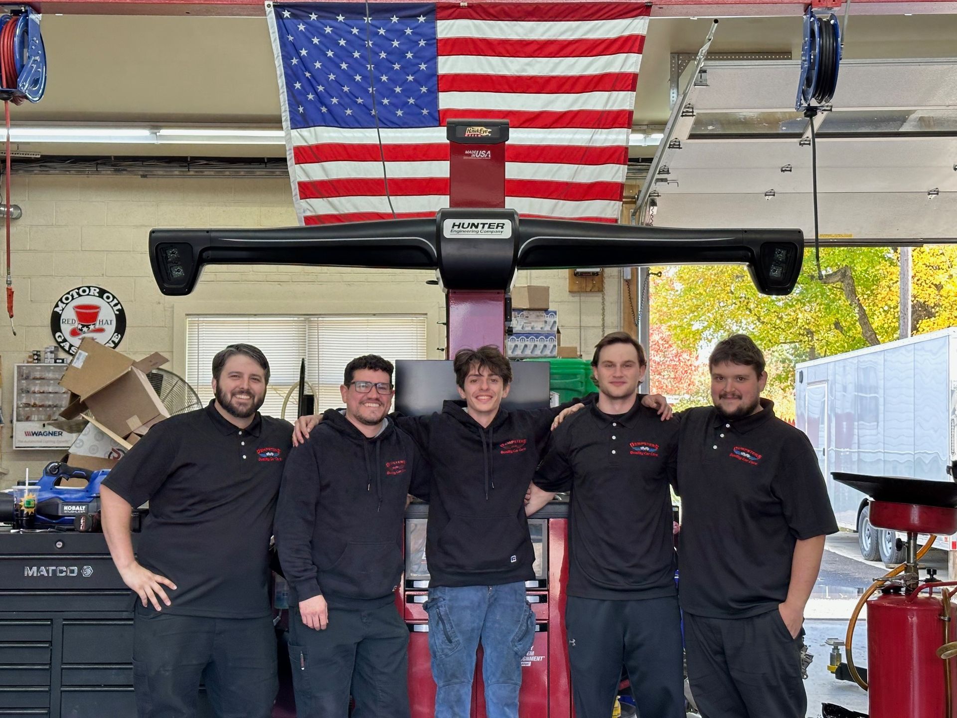 Five people standing together in an auto repair shop with an American flag hanging in the background.
