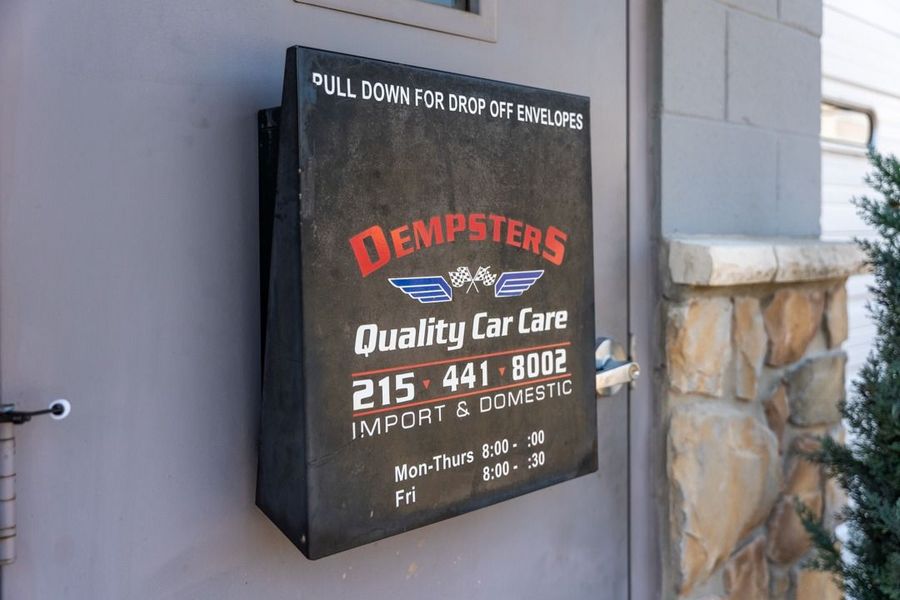 Black sign for Dempster's Quality Car Care on a gray wall. Includes a phone number and hours of operation.