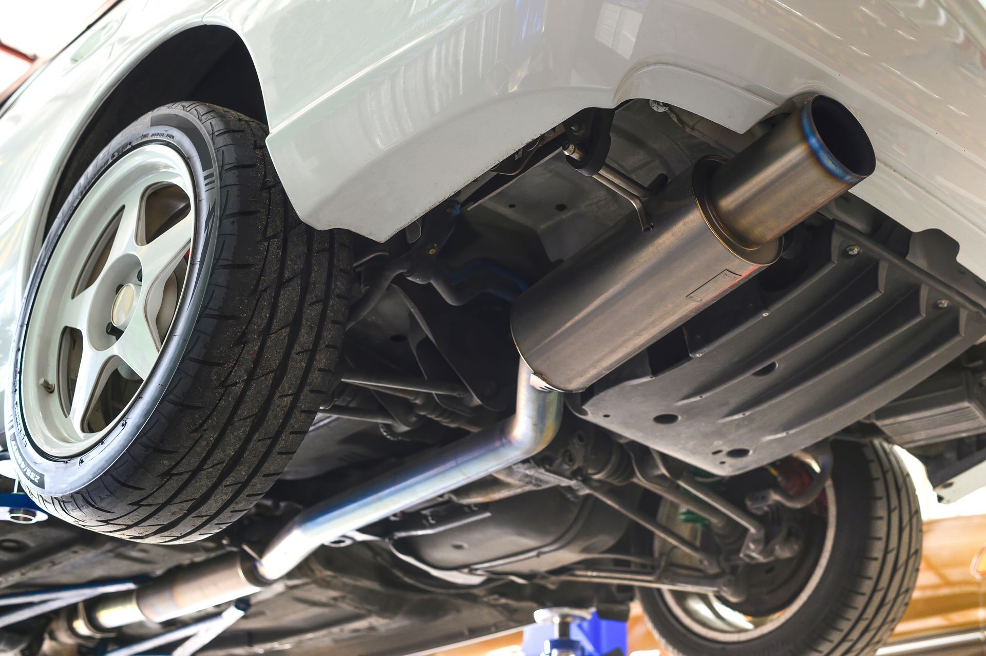 Underside of a white car with its exhaust system on display. Shows a wheel, pipes, and muffler.