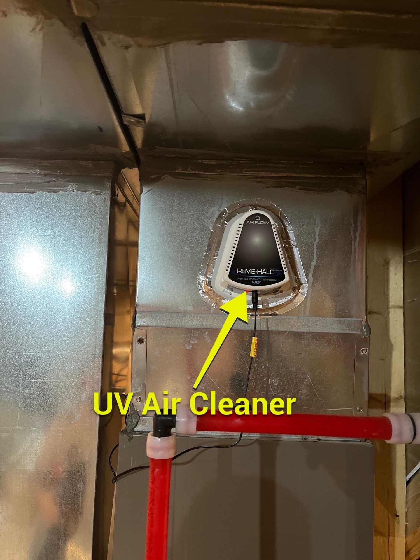 A UV air cleaner unit mounted on a silver metal HVAC duct, with a yellow arrow pointing to the device.