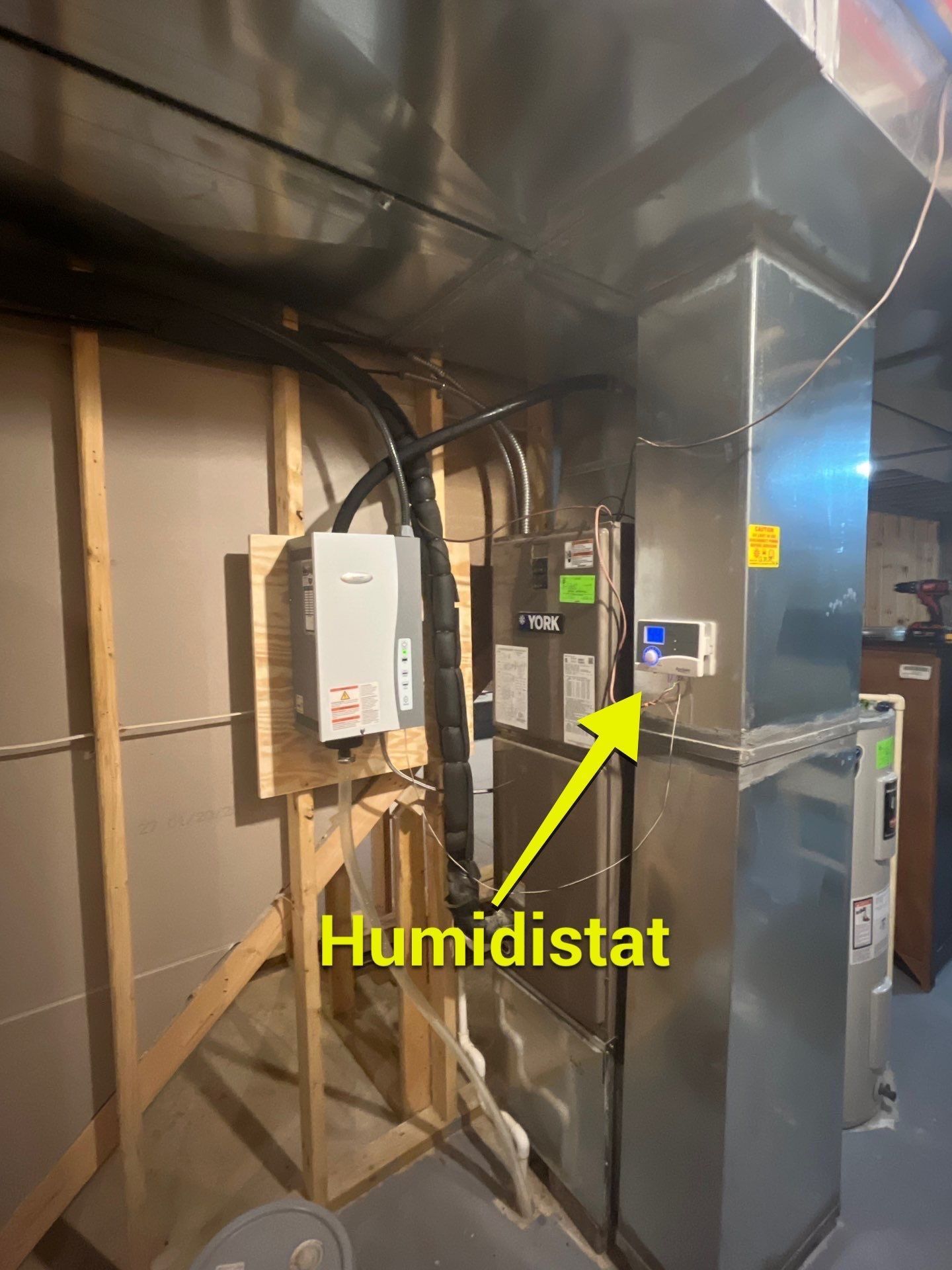 A furnace in a basement with a wall-mounted humidistat highlighted by a yellow arrow and label.