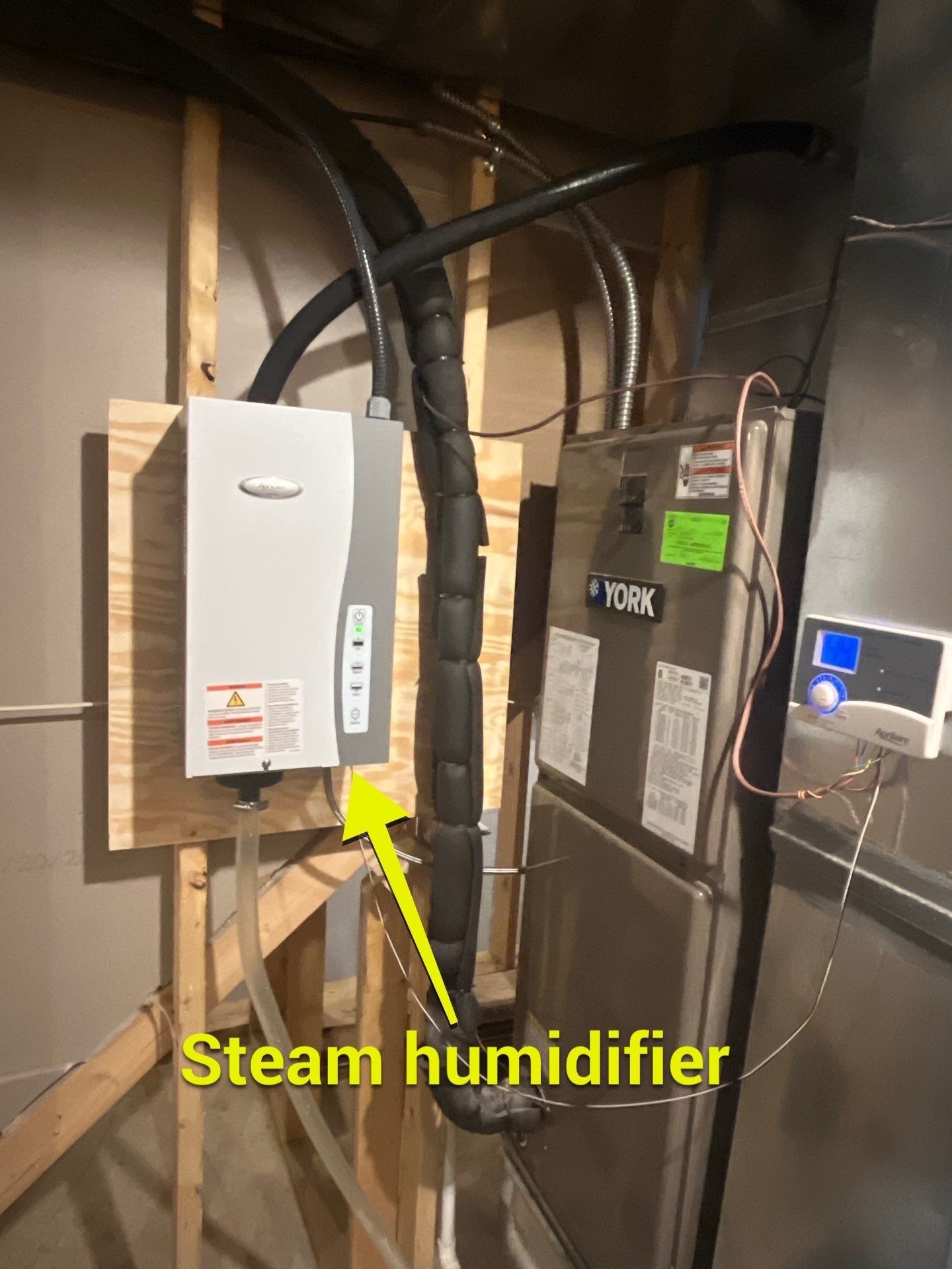 A steam humidifier mounted on a wooden board next to a furnace unit, connected by a large, insulated flexible hose.