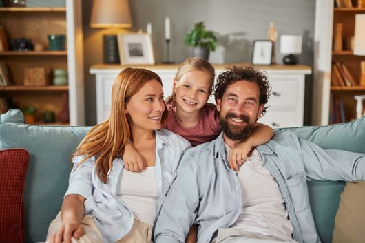 A family of three sitting on a light blue sofa in a living room, smiling at the camera.