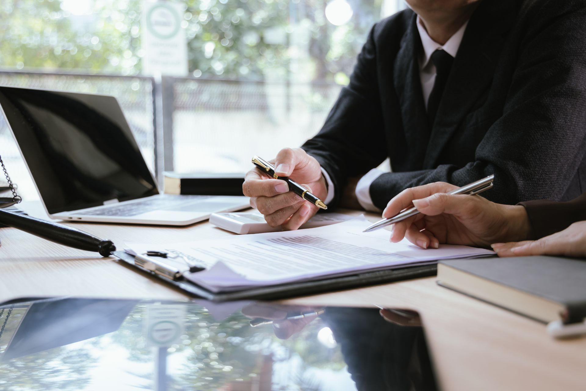 Two professionals in business attire review a document together at a desk with a laptop and a clipboard.