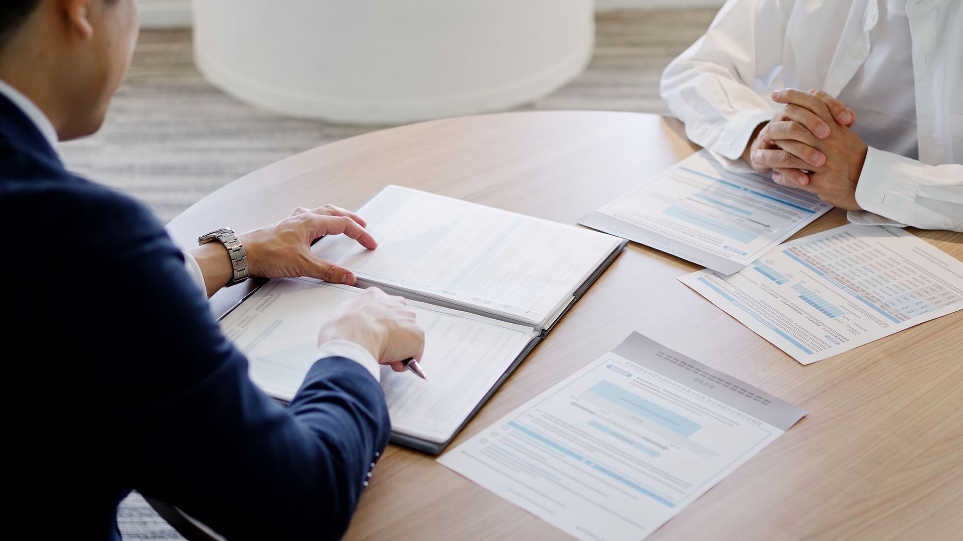 Two professionals in business attire review documents and charts at a circular table in an office setting.