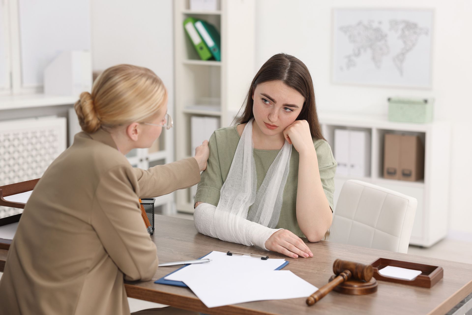 A person comforts another person with a bandaged arm at a desk featuring a gavel and documents in an office setting.