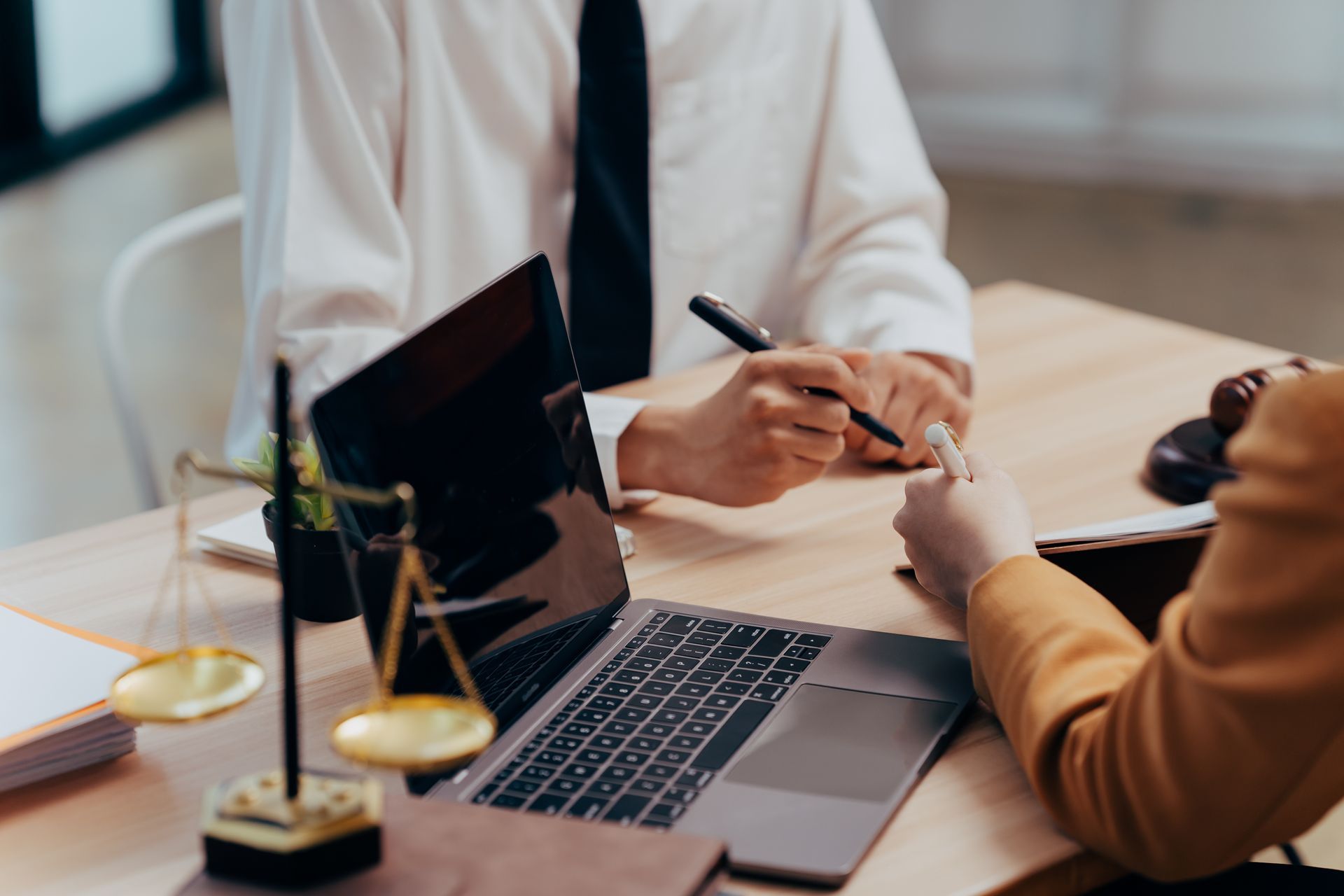 Two professionals sit at a desk with a laptop and scales of justice, discussing a document during a meeting. Two professionals sit at a desk with a laptop and scales of justice, discussing a document during a meeting.