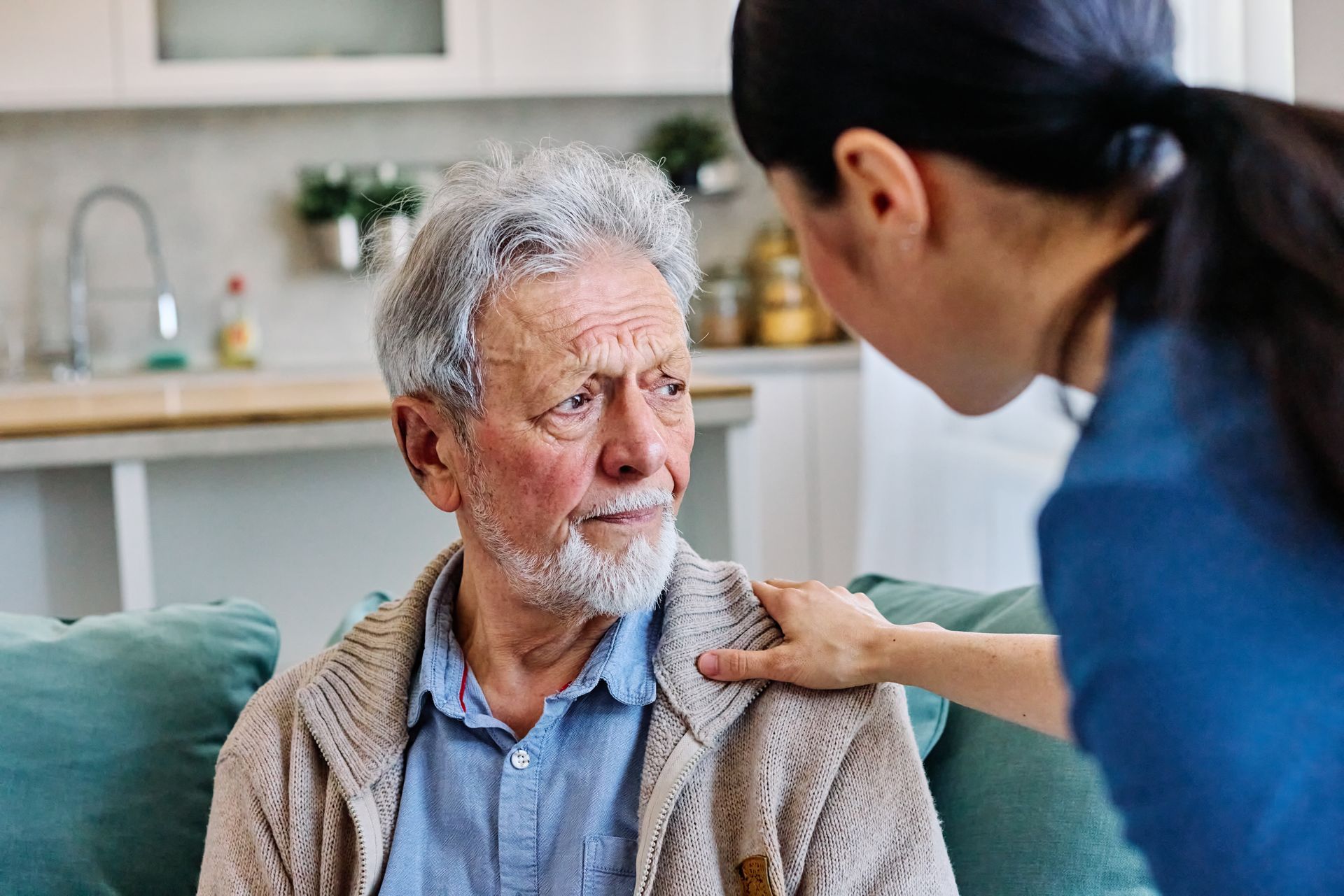 A caregiver with their hand on an elderly person's shoulder as they look at each other in a softly lit kitchen setting.