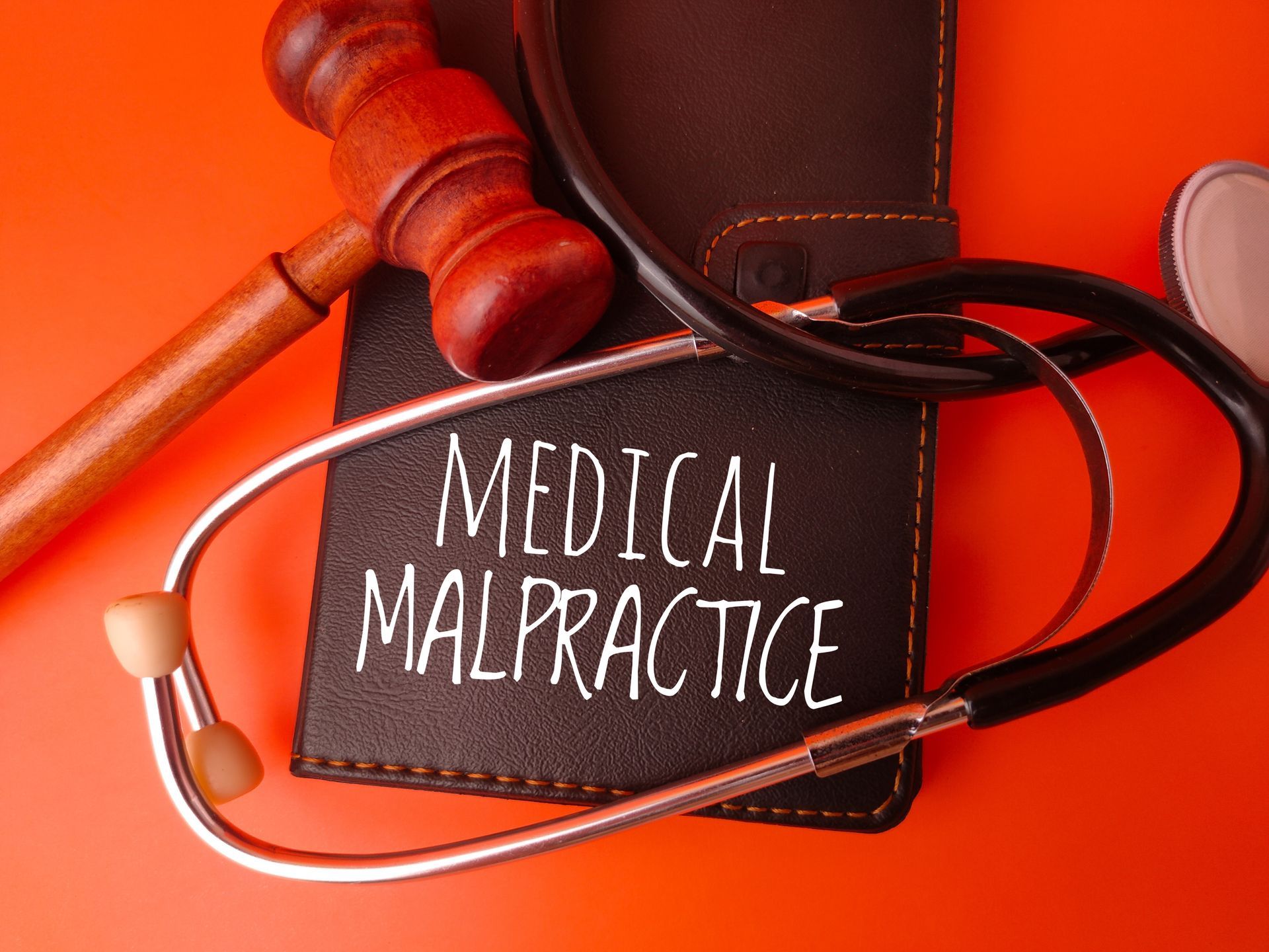 A wooden gavel and a stethoscope rest on a dark notebook labeled MEDICAL MALPRACTICE against an orange background.