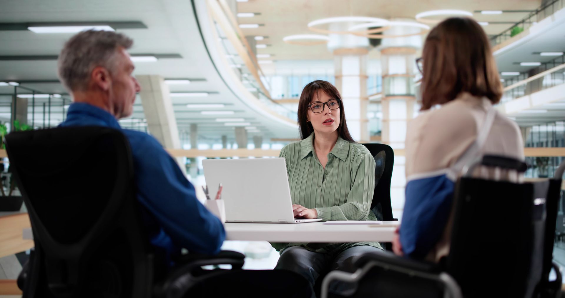 Three people sitting around a desk in a modern office space, engaged in a discussion with an open laptop between them.