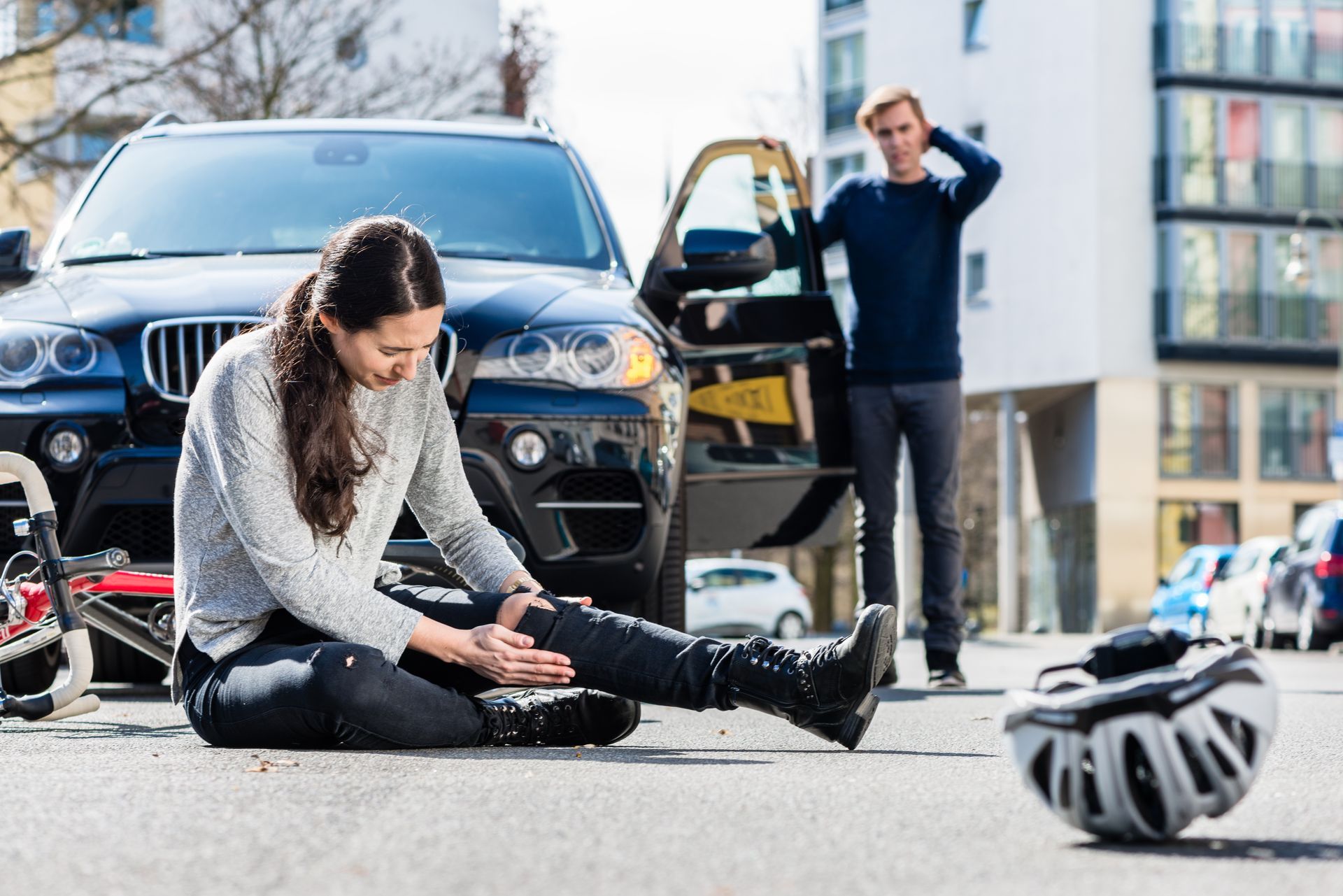 A person sits on the asphalt clutching their injured leg after a bike accident near a car; another person looks on.