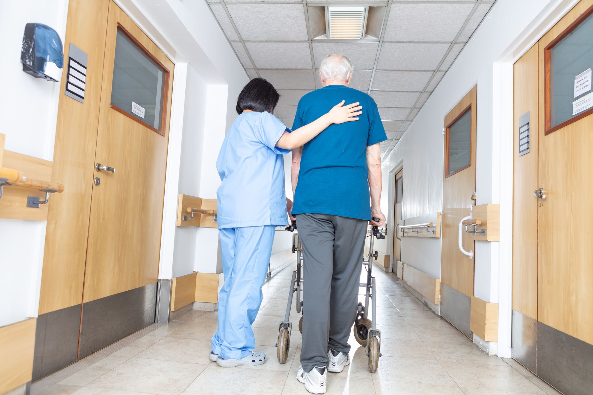 A caregiver in blue scrubs assists a person using a wheeled walker down a brightly lit hospital or care facility hallway.