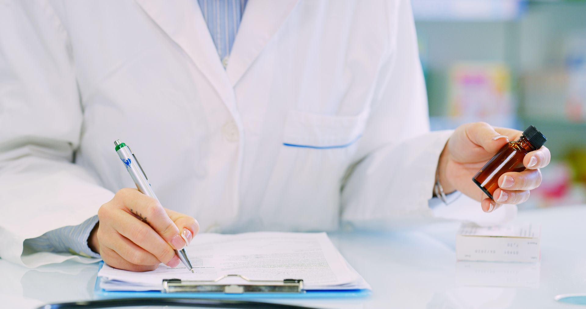 A person in a white lab coat writing on a clipboard while holding a small amber medicine bottle.