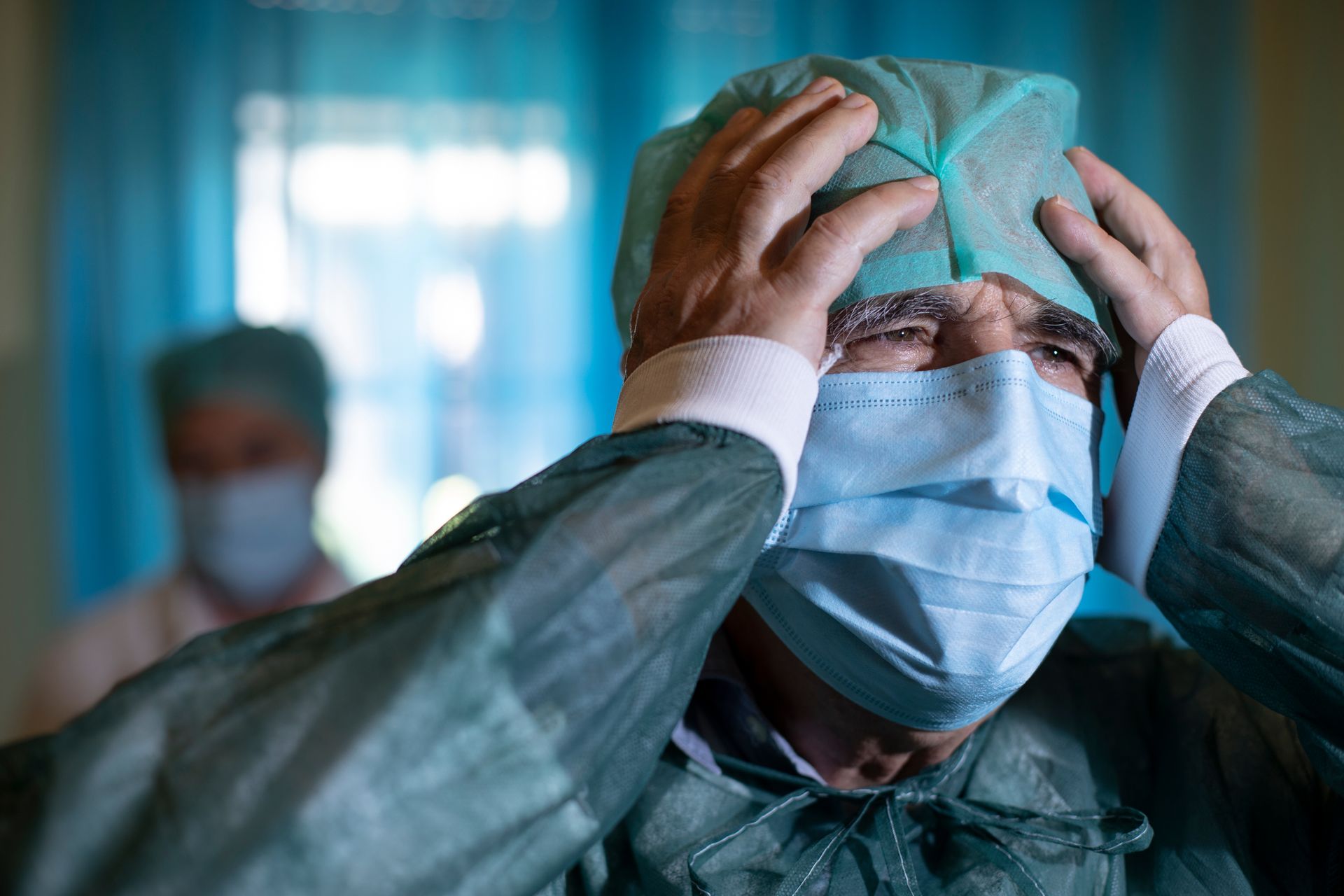 A person in medical scrubs, a surgical cap, and a face mask holding their head with both hands in a distressed manner.