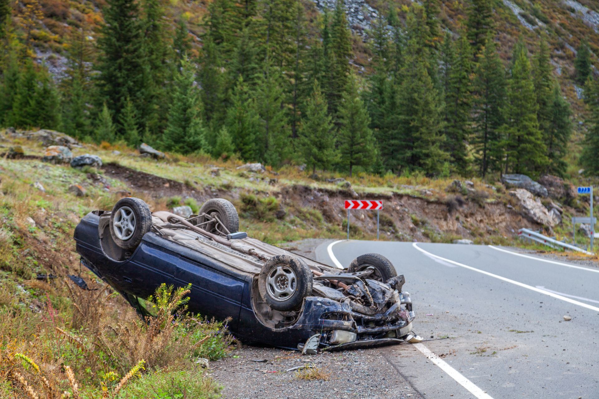 A dark blue car lies overturned on the side of a mountainous road near a forest.
