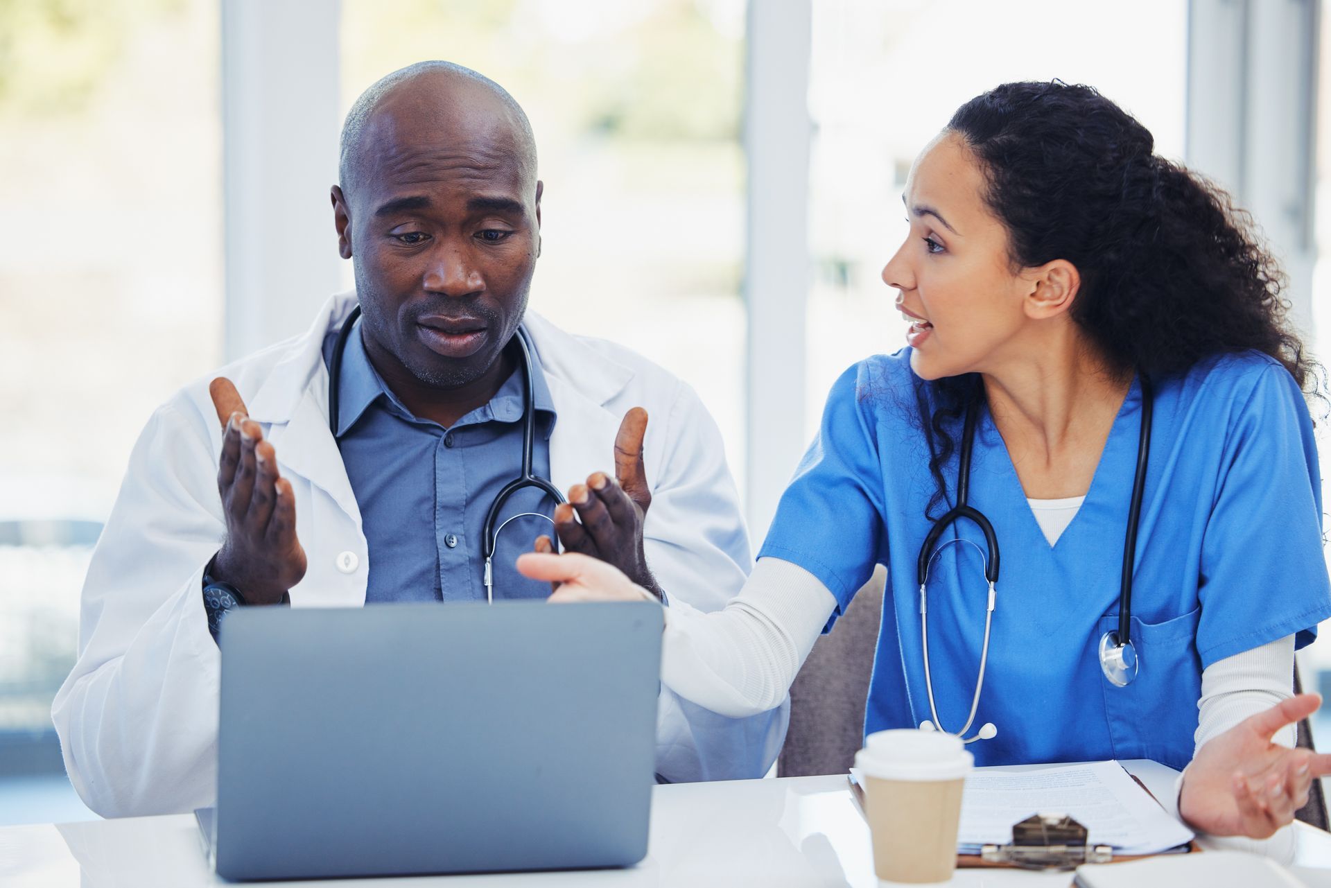 Two healthcare professionals in scrubs and lab coats sit at a desk, looking at a laptop and gesturing during a discussion.
