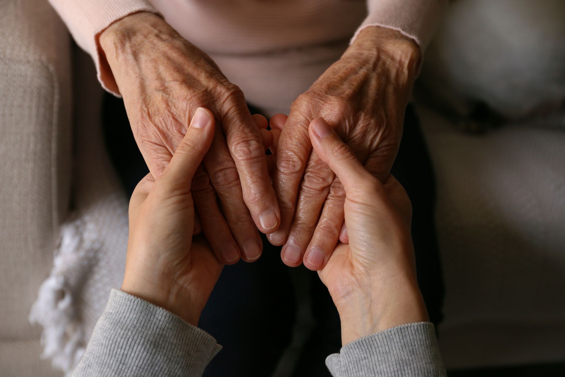 Close-up, high-angle view of two hands holding the weathered, wrinkled hands of another person in a gesture of support.