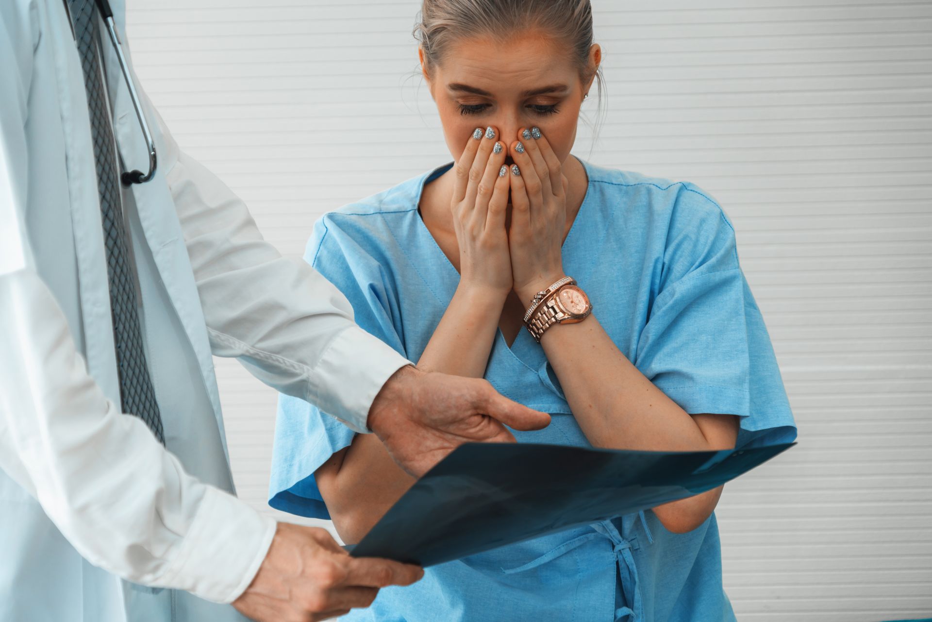 A doctor shows an X-ray to a patient in scrubs who looks upset with their hands held to their face.