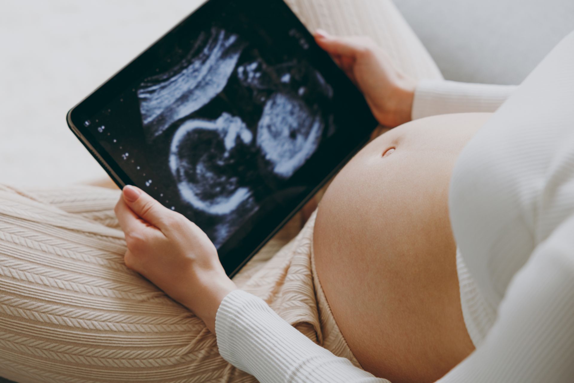 A pregnant person in light-colored clothing holds a tablet displaying an ultrasound scan of a fetus.