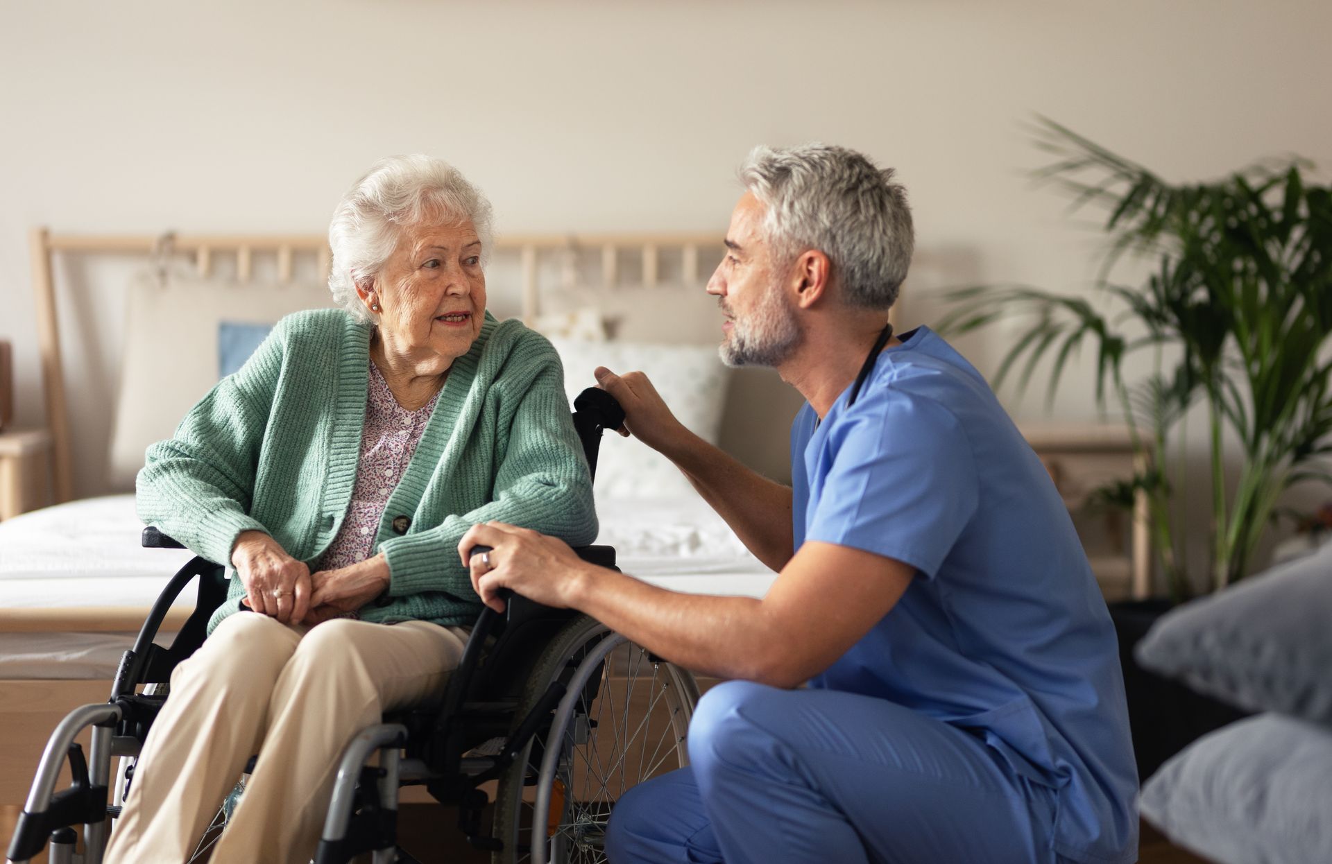 A healthcare worker in blue scrubs kneels beside an individual sitting in a wheelchair to speak with them in a bedroom.