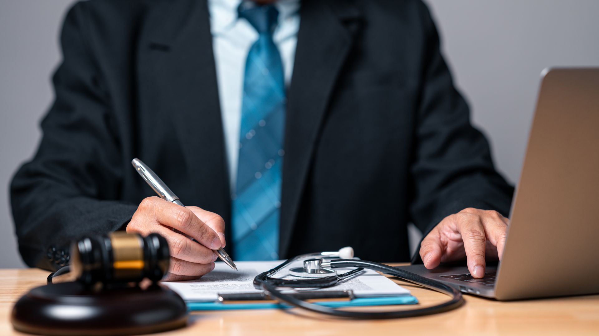 A person in a suit writes on a document next to a gavel, stethoscope, and laptop on a desk.