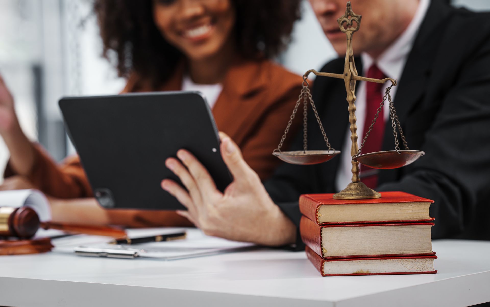 Two professionals look at a tablet near a gavel and a set of law books with a scale of justice on top.