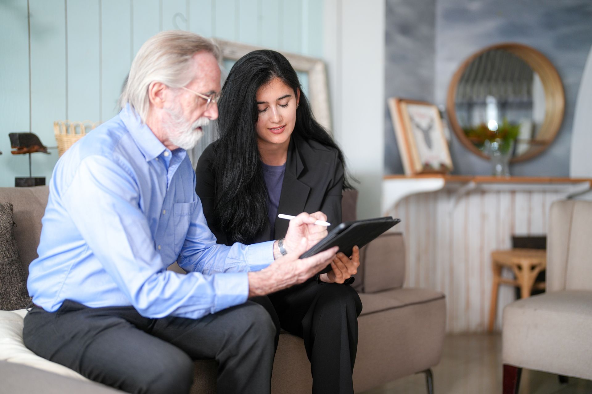 A person in a blue shirt and another in a black blazer sit together on a sofa, reviewing a digital tablet.