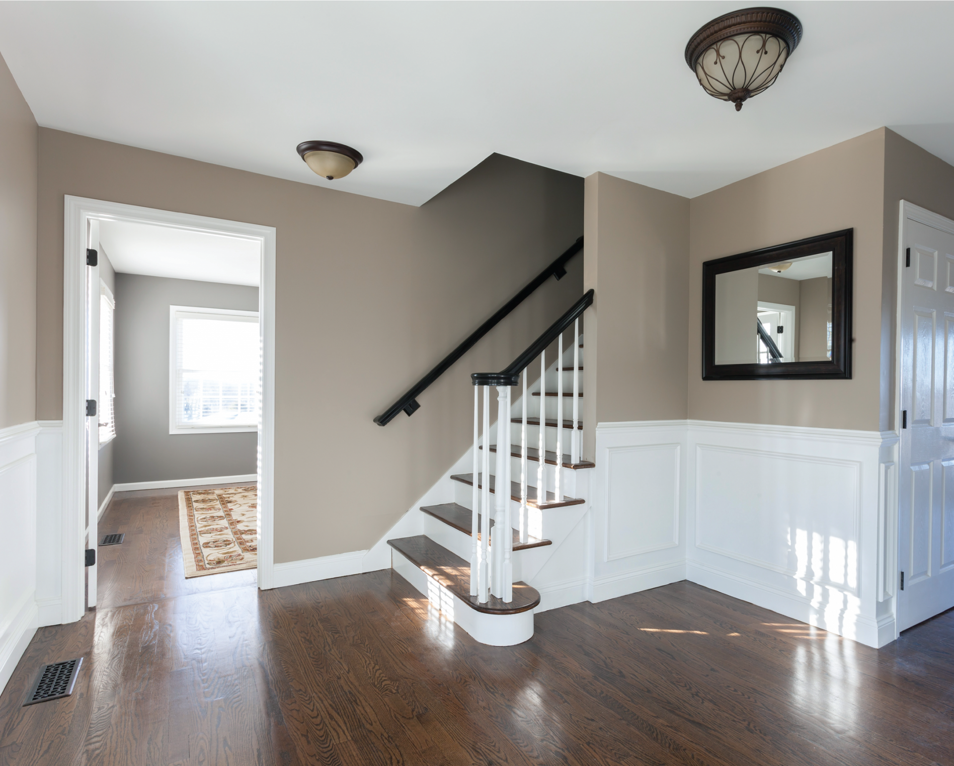 Entryway with staircase, dark wood floors, white wainscoting, beige walls, mirror, and two lights.