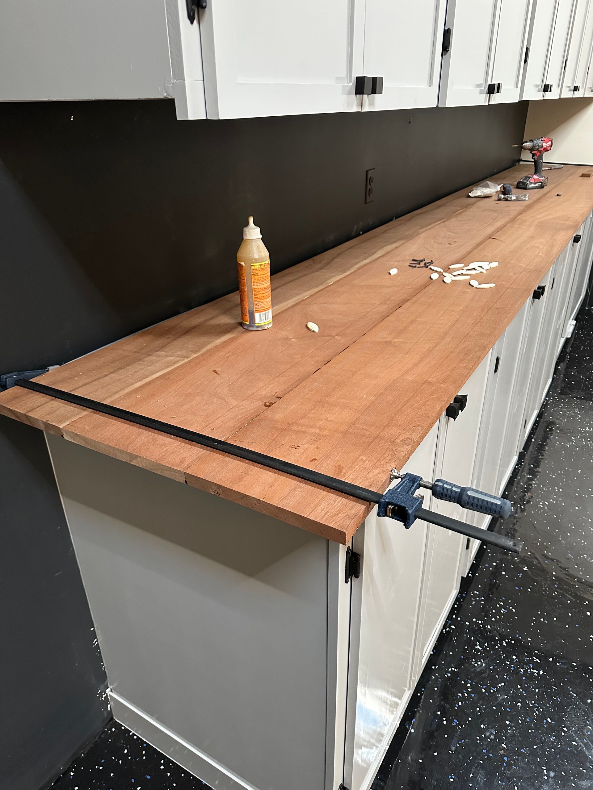 Wooden workbench being assembled on white cabinets, clamped in place. Brown glue bottle on top.