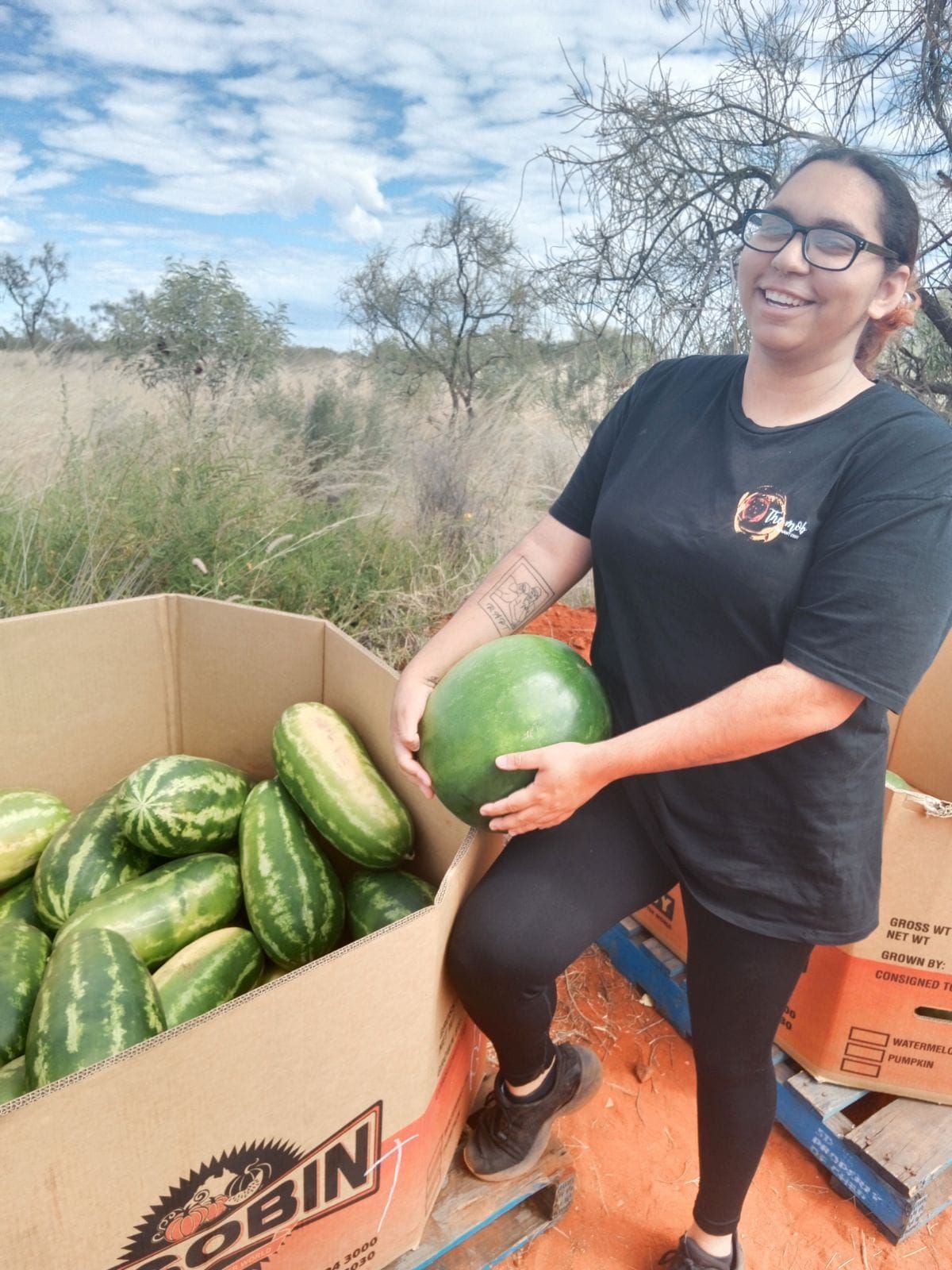 Woman smiling, holding a watermelon, near a box of watermelons on a pallet outdoors.