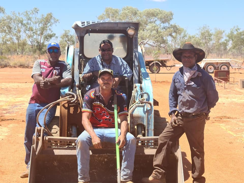 Four men pose in front of and in a blue tractor outdoors on red earth. One man sits, others stand. They are smiling.