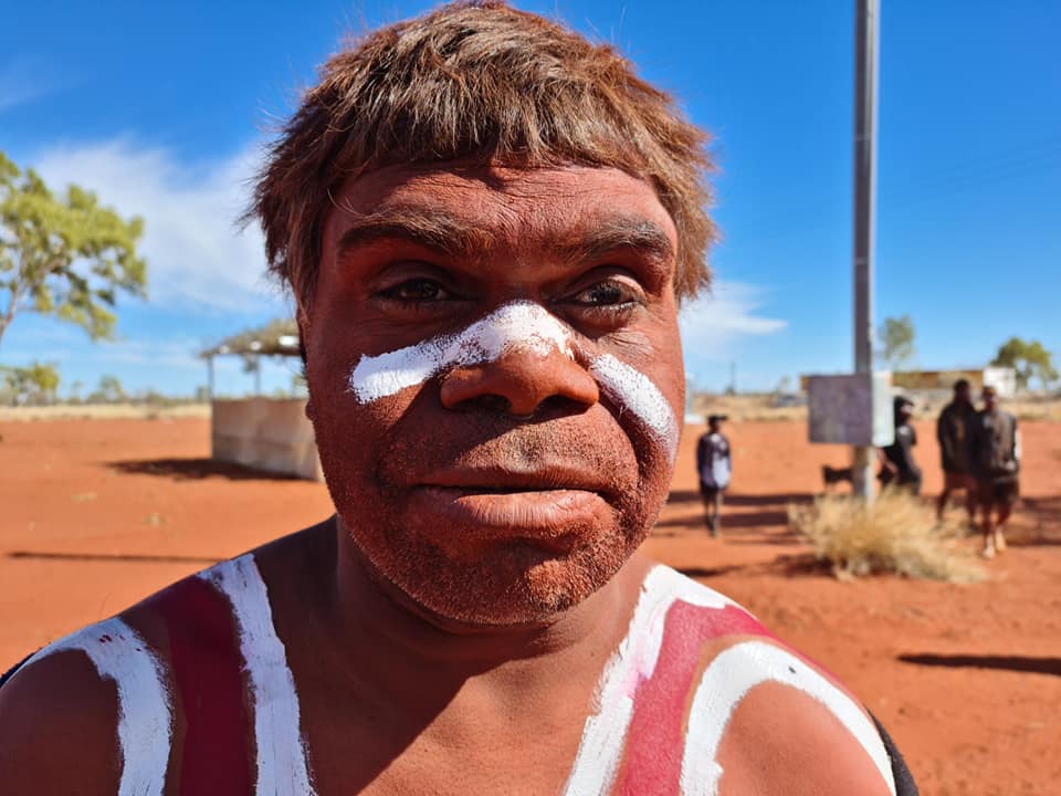 Indigenous Australian man with red body paint and white face markings, in an outdoor setting with others.