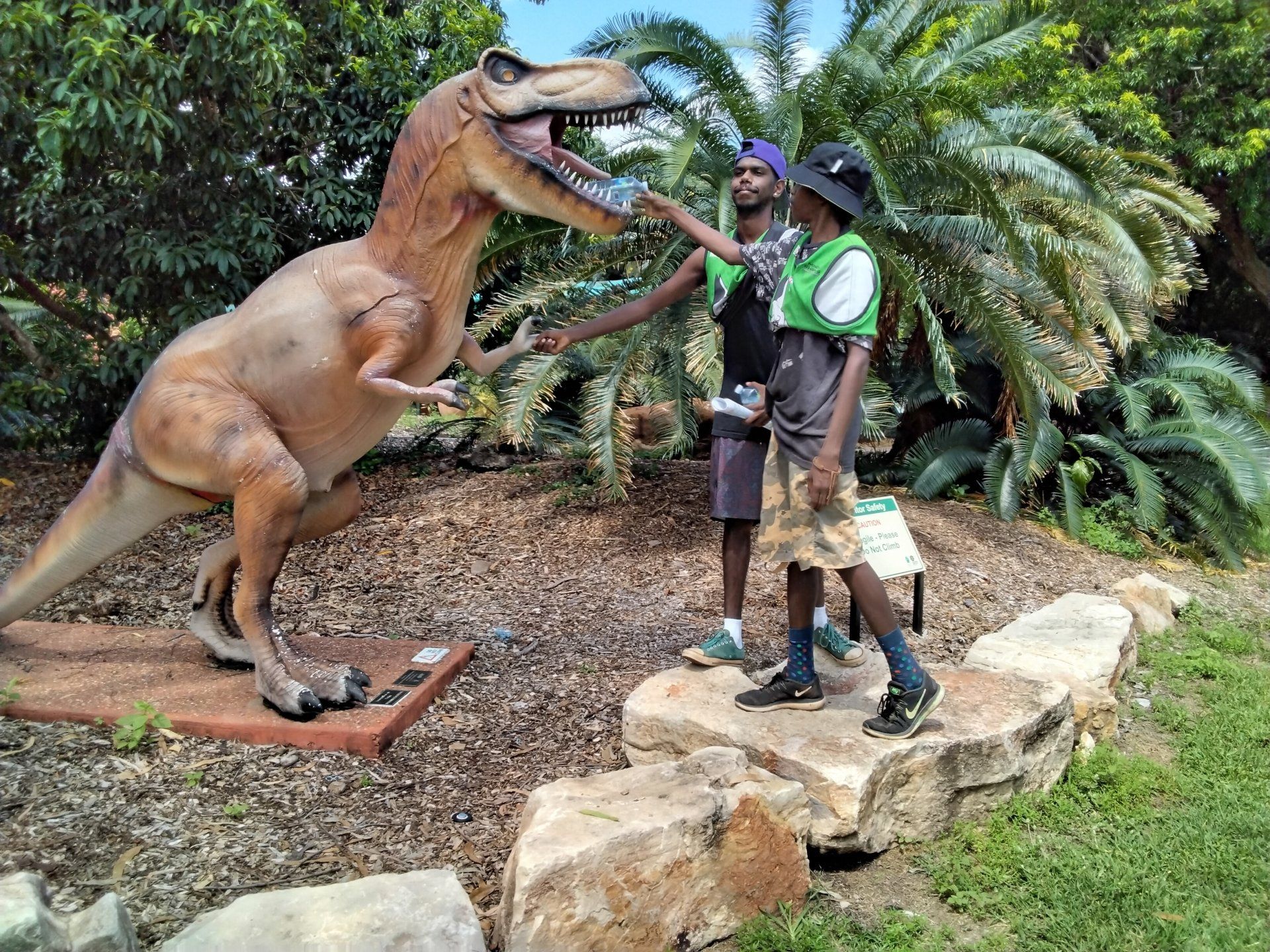 Two people reaching toward a T-Rex statue in a garden setting, under a blue sky.