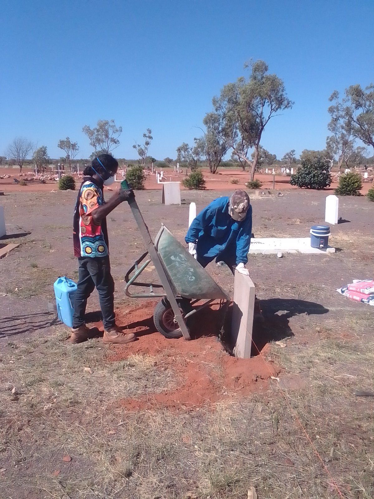 Two people working at a cemetery: one tipping a wheelbarrow near a tombstone.