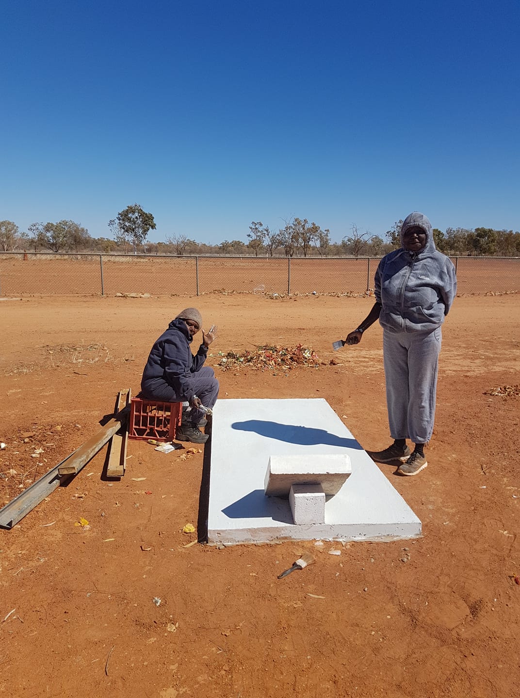 Two people, one sitting and one standing, beside a freshly painted cement structure in a dusty, rural setting.