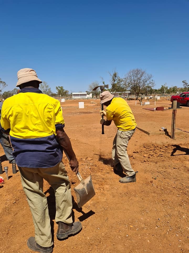 Two people in yellow shirts and hats digging in red dirt under a blue sky. They appear to be working on a construction site.