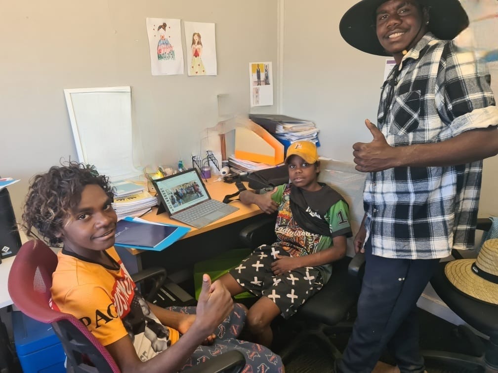 Three Indigenous Australians in an office setting; two seated, one standing. The standing man gives a thumbs-up.