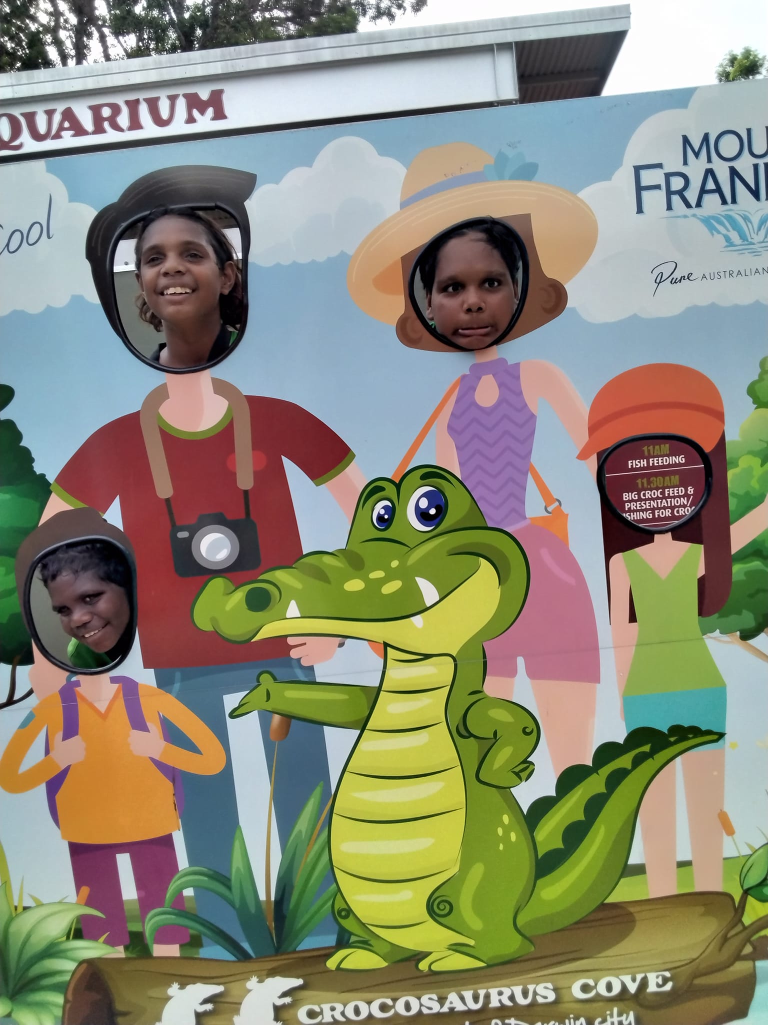 People posing in cutouts at a Chocosaurus Cove photo op. Features a crocodile and several characters with face holes.