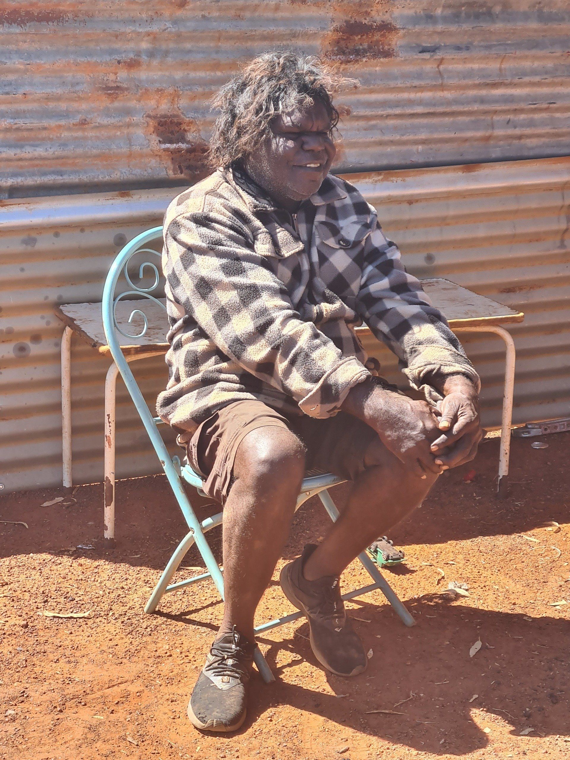 An Indigenous Australian man sits outside, smiling. He wears a plaid shirt and shorts, and rests his hands on his lap.