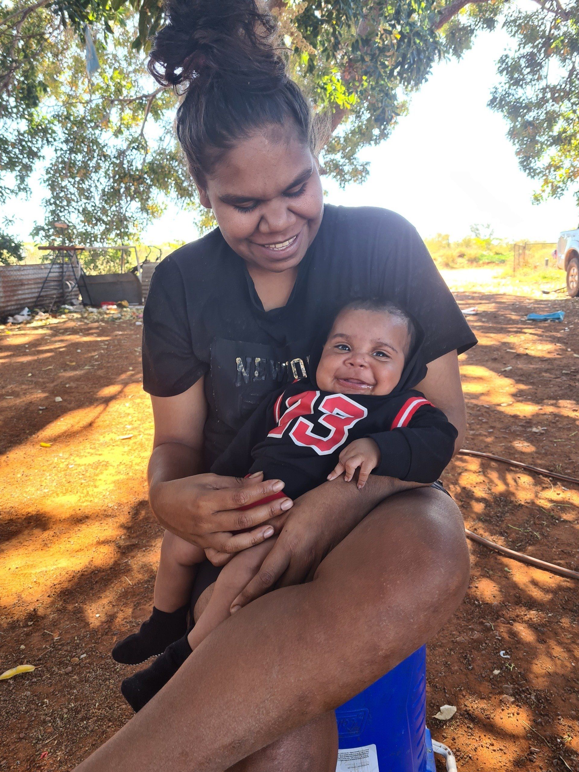 Woman holding a smiling baby, both outside. The baby wears a black and red sweatshirt. They are sitting under a tree.