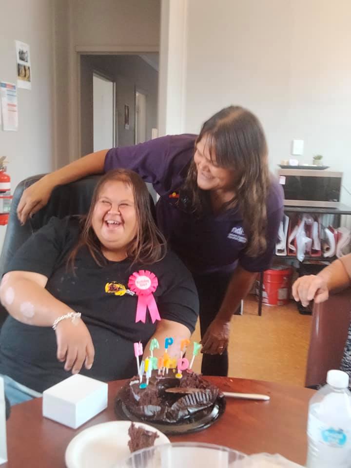 Two women celebrating a birthday. One seated, laughing, with a birthday pin and cake. The other leans in, smiling.