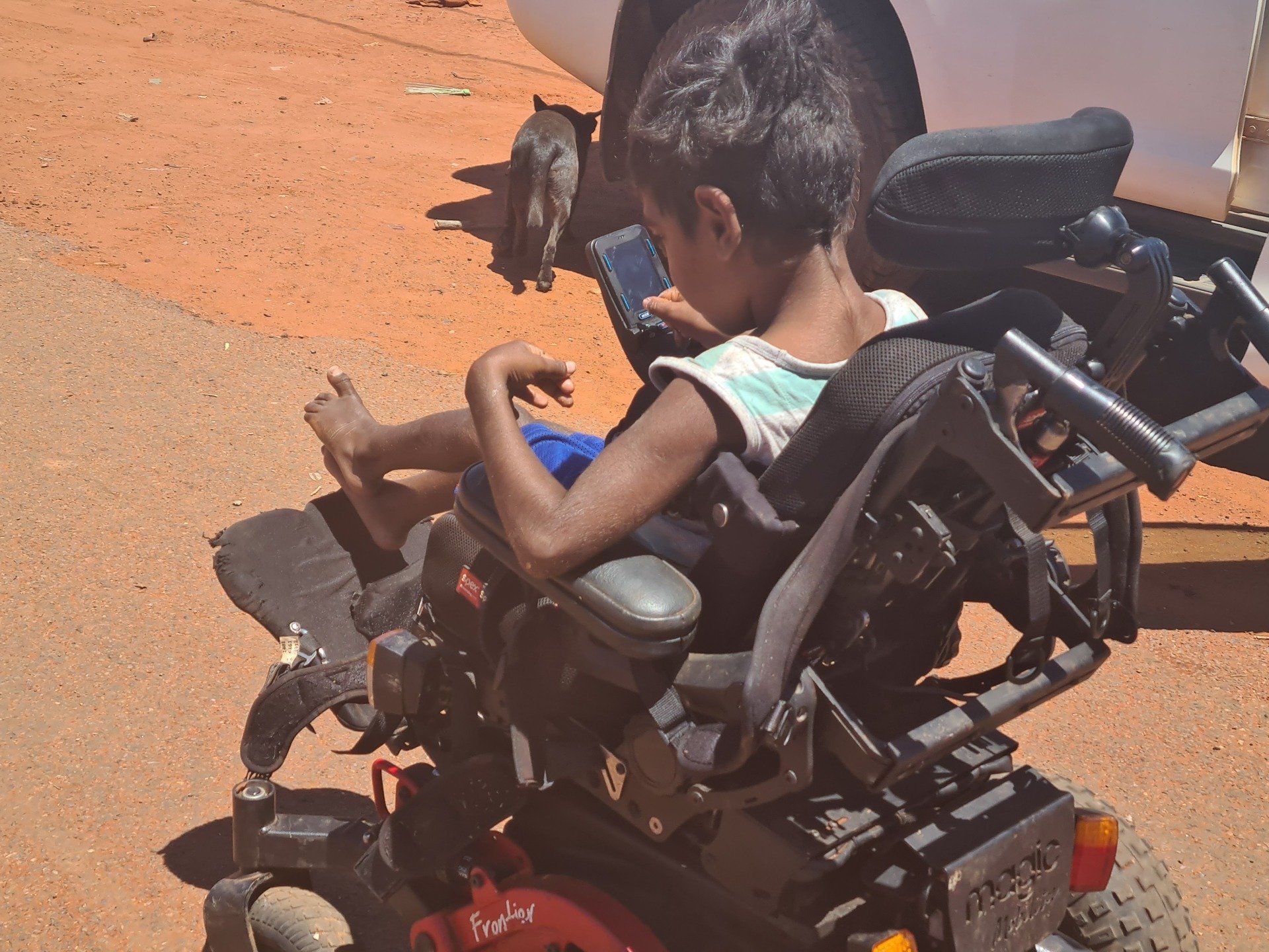 Child in a motorized wheelchair, using a tablet outdoors on red dirt. A small dog is nearby.